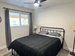 Ein Schlafzimmer mit einem Bett, einem Nachttisch, einer Lampe, einem Fenster mit Vorhängen und einem Deckenventilator.