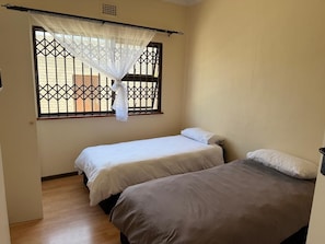 A room with two beds, a window with a metal grille, and a curtain. - Rihla Residences - Hanbury (Cape Town)