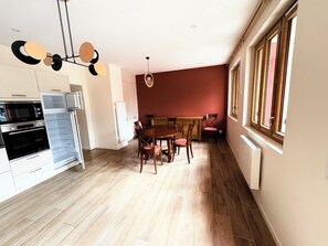 A modern kitchen with built-in appliances, a dining area with a wooden table and chairs, and a large window with blinds. - Downtown ground-floor apartment (La Chapelle-en-Vercors)