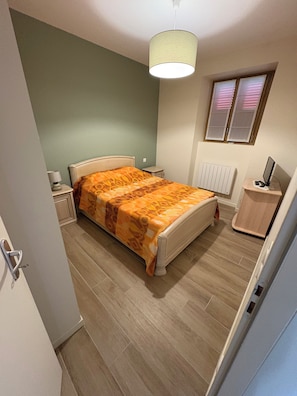 Une chambre à coucher comprenant un lit, une table de chevet, une lampe, un radiateur, une fenêtre avec stores et un bureau avec un ordinateur.