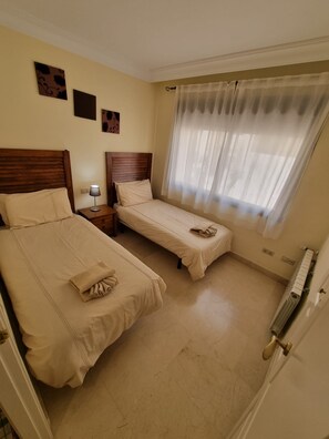 Une chambre d’hôtel avec deux lits, une table de chevet, une lampe, une fenêtre avec des rideaux légers et des œuvres d’art murales.