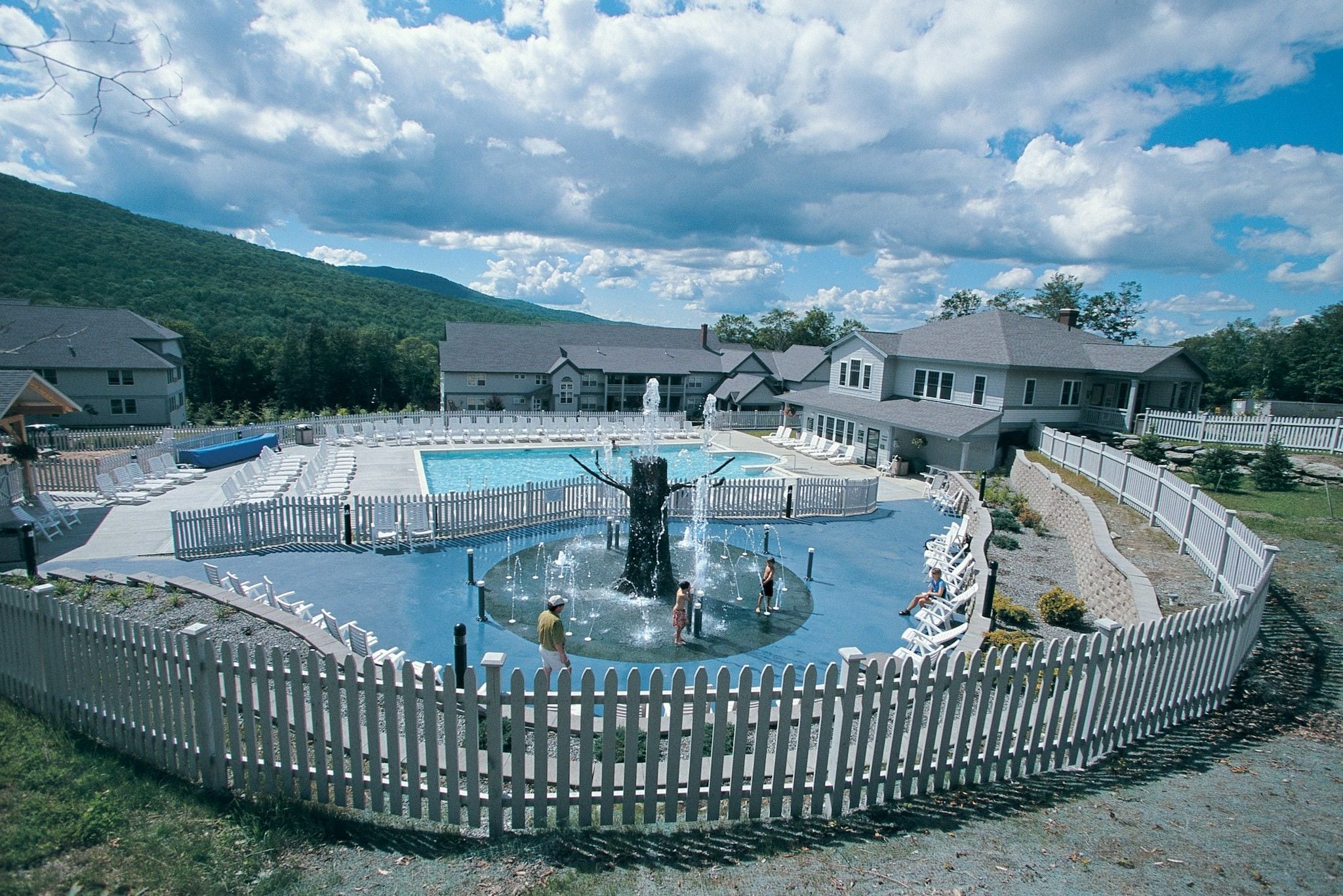 A swimming pool with a central fountain, surrounded by a white picket fence, and adjacent to a building with a porch.