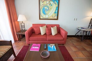A red sofa with two pillows, a coffee table with magazines and a remote control, a painting on the wall, and a lamp on a side table. - Jr Suite at The Club at Hacienda del Mar (Cabo San Lucas)