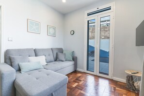 A living room with a grey sofa, a small ottoman, a clock, and a door leading to an outdoor area. - Chez Agostinha by Atlantic Holiday (Funchal)