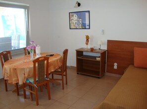Een kamer met een eettafel, stoelen, een bed, een nachtkastje, een lamp, een foto aan de muur en een raam met jaloezieën.