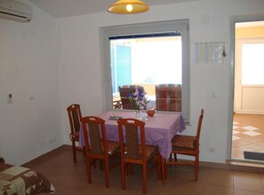 A dining area with a table set for four, surrounded by chairs, a window with blinds, and a view of a balcony. - One Bedroom Apartment with Balcony and Sea View (Petrcane)
