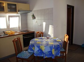 A compact kitchen with a dining table covered by a blue tablecloth, wooden chairs, a stove, and a window providing natural light. - One Bedroom Apartment with Terrace and Garden (Petrcane)