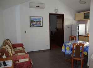 Um espaço de estar compacto com sofá, mesa de jantar com cadeiras, cozinha com geladeira e porta que dá para outro cômodo.