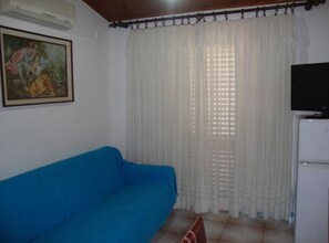 A room with a blue sofa, a television on a stand, and a window with white curtains. - One Bedroom Apartment with Terrace and Sea View (Petrcane)