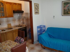 A compact kitchen with wooden cabinets, a stove, and a sink. There is a dining area with a table and chairs. A blue sofa is against the wall. - One Bedroom Apartment with Terrace and Sea View (Petrcane)