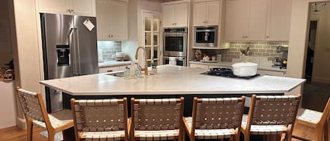 A kitchen with a large island, white cabinets, stainless steel appliances, and wooden chairs.