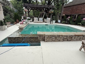 A swimming pool with a stone border, surrounded by outdoor furniture and a pergola with hanging lights. - Lovely 4-bedroom house with AC in pleasant Tulsa neighborhood (Tulsa)