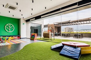 Ein Kinderspielbereich mit grüner Wand, bunten Spielgeräten und Blick auf eine Stadtlandschaft durch große Fenster.