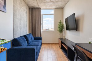 Ein modernes Wohnzimmer mit einem blauen Sofa, einem an der Wand montierten Fernseher und einem Fenster mit Blick auf die Stadt.