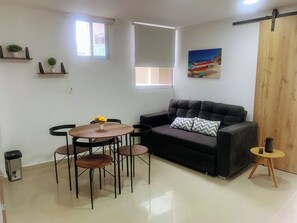 Ein Wohnzimmer mit einer Couch, einem runden Tisch mit Stühlen, einem kleinen Mülleimer und einem Fenster mit Jalousien.