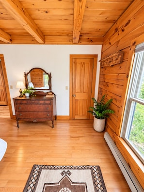 A wooden interior with a bed, dresser, mirror, and a window with a view. - Handcrafted Log Home on North Haven Island, Maine (North Haven)