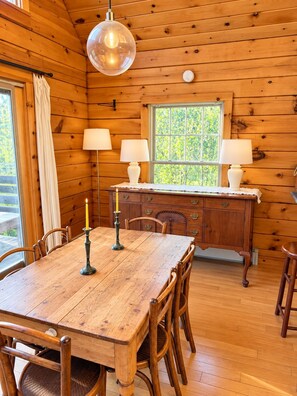 A wooden cabin interior with a dining table, chairs, a chandelier, and a window with a view of greenery. - Handcrafted Log Home on North Haven Island, Maine (North Haven)