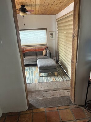 A room with a grey sofa, a small ottoman, a ceiling fan, a window with blinds, and a picture on the wall. - Lovely 2-bedroom house in tranquil Taos. (Taos)