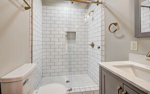 A bathroom with white subway tiles, a toilet, a shower with a glass door, and a vanity with a mirror. - Charming 6BR Norman Home Perfectly Near OU and Campus Corner (Norman)
