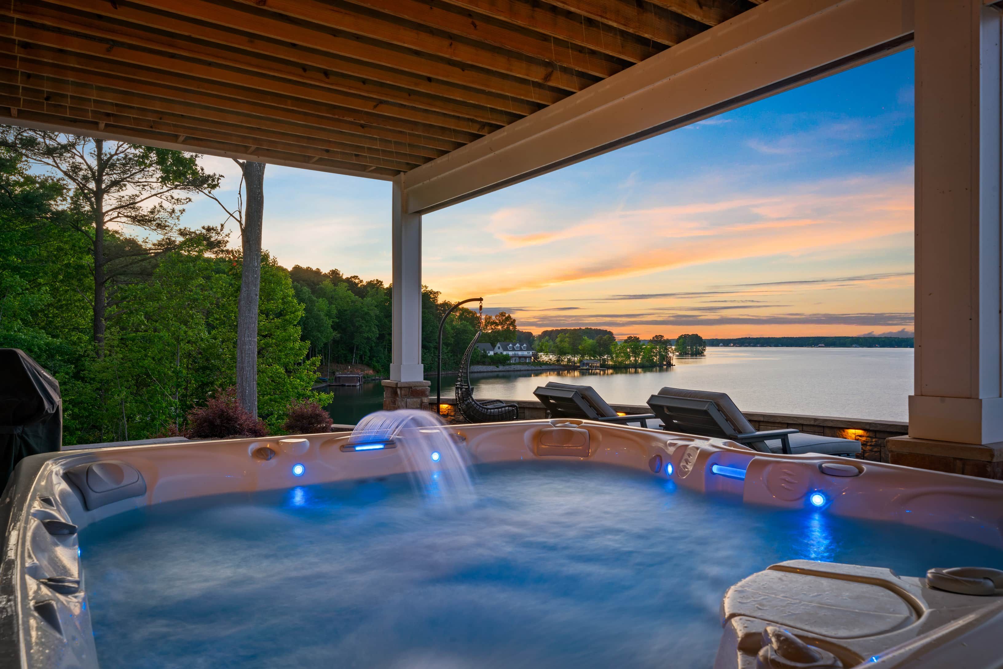 Un jacuzzi con caída de agua, con vista a un lago y al atardecer, además de un patio cubierto con sillones de descanso.