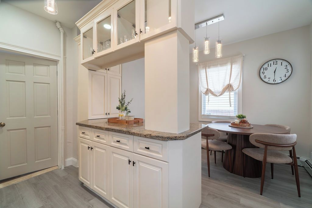 A kitchen with white cabinets, a granite countertop, a dining area with a wooden table and chairs, and a wall clock.