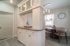 Una cocina con armarios blancos, encimera de granito, comedor con mesa y sillas de madera, y un reloj de pared.