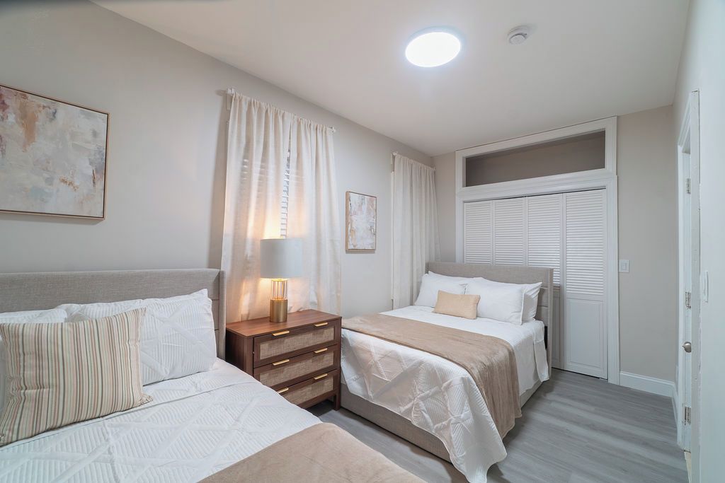 Una camera d'albergo con due letti, un comodino con una lampada, una finestra con tende e una porta che dà su un panorama.