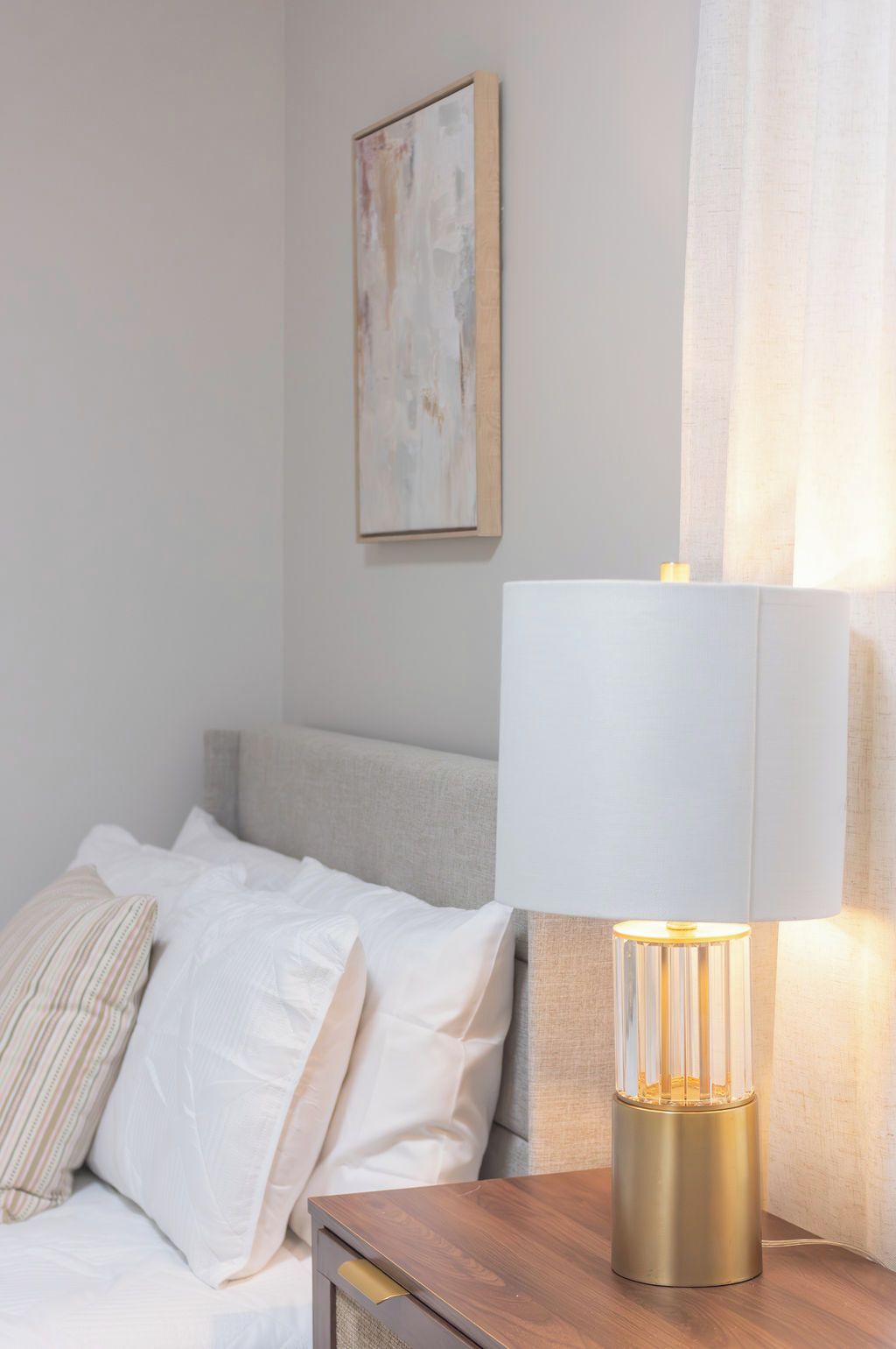 A neatly made bed with white linens and a single striped pillow, a bedside table with a lamp, and a painting on the wall.