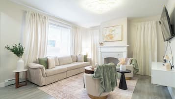 Ein Wohnzimmer mit einem beigen Sofa, einem runden Couchtisch, einem Kamin und einem an der Wand montierten Fernseher.