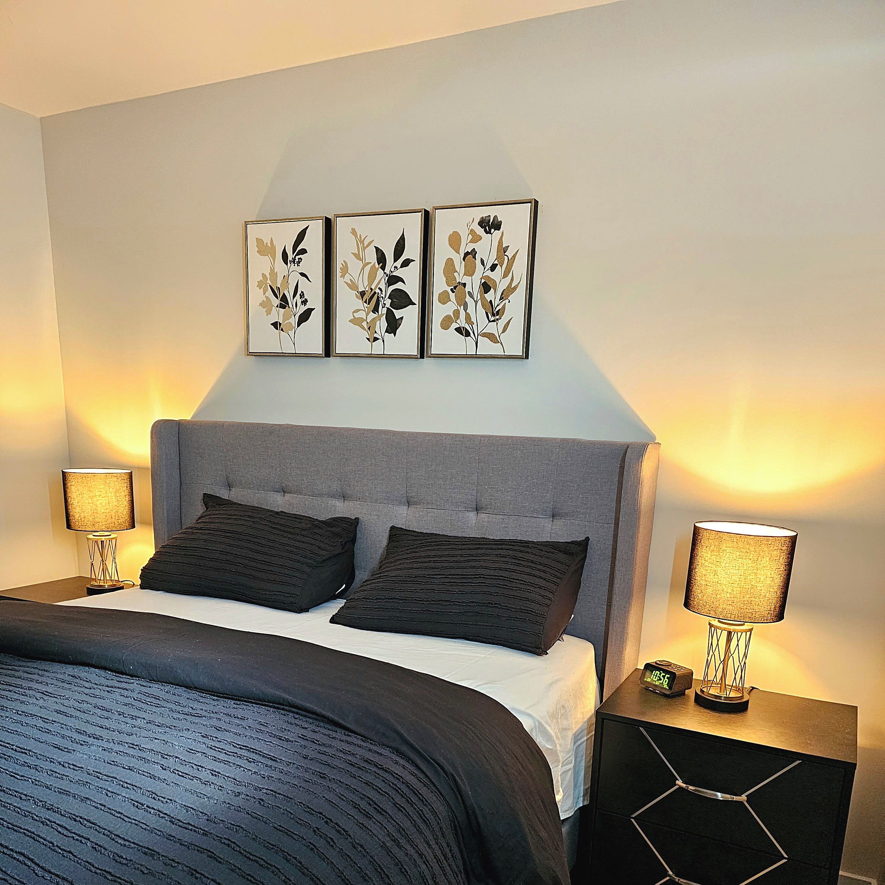 A bedroom with a grey upholstered headboard, a bed with dark bedding, two bedside lamps, and three framed botanical prints on the wall.