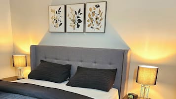 A bedroom with a grey upholstered headboard, a bed with dark bedding, two bedside lamps, and three framed botanical prints on the wall.