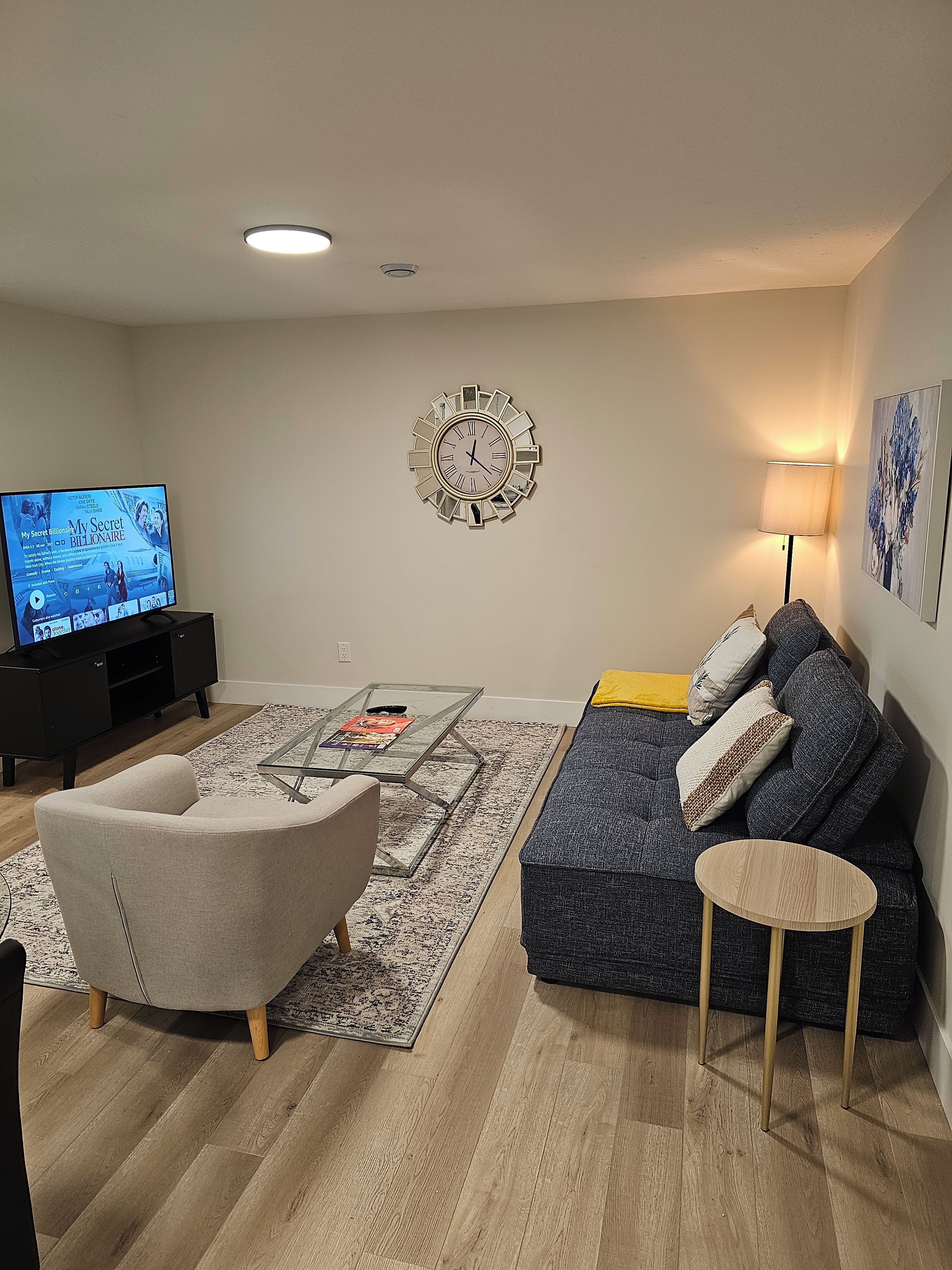 Uma sala de estar com televisão, sofá, poltrona, mesa de centro e relógio decorativo na parede.