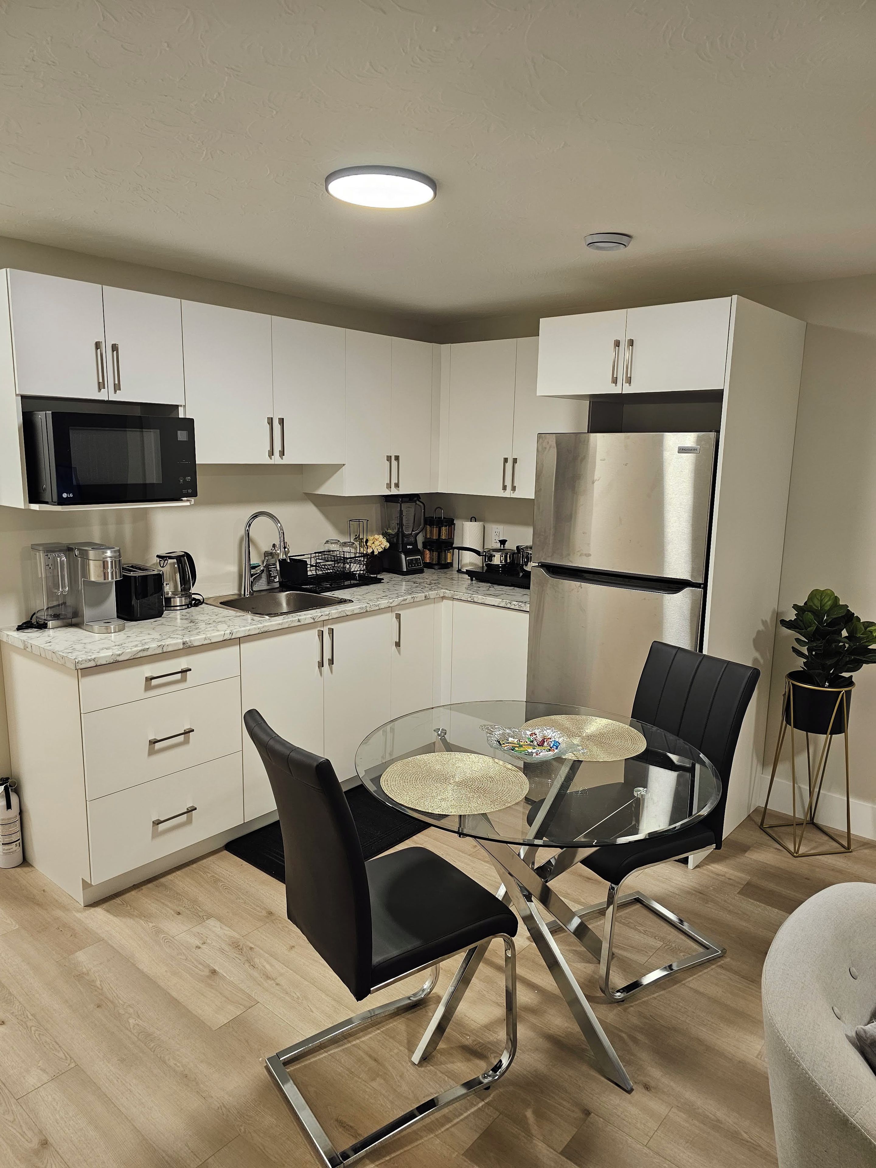 A modern kitchen with white cabinets, a black microwave, a stainless steel refrigerator, and a glass dining table with black chairs.