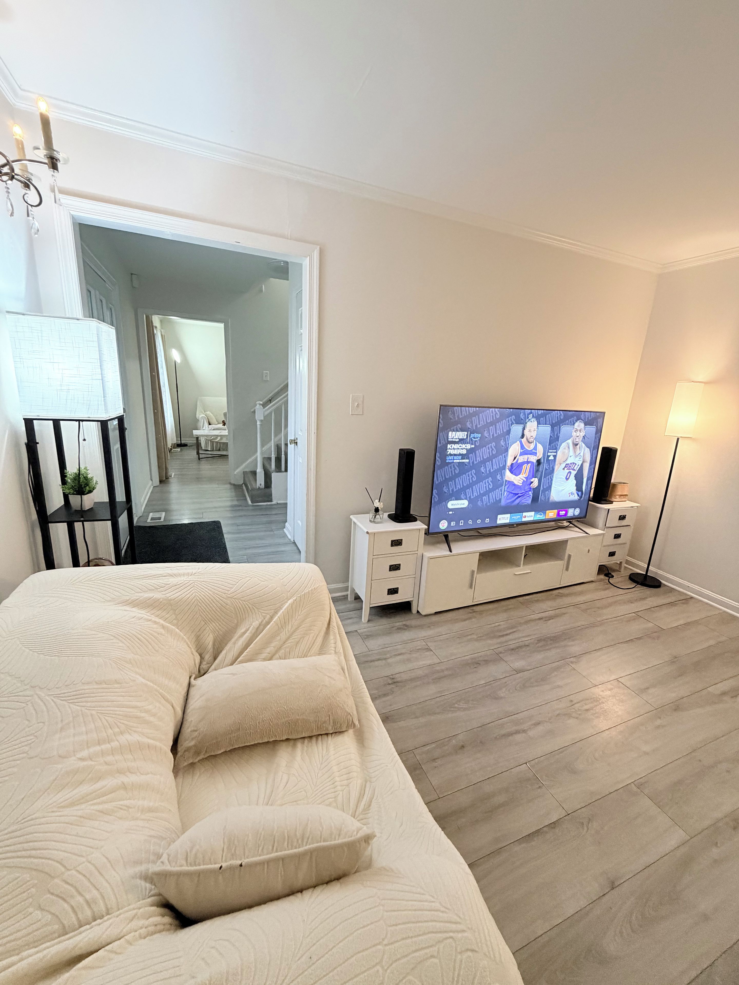 Een woonkamer met een beige bank, een flatscreen televisie op een standaard, een vloerlamp en een trap die naar een andere kamer leidt.