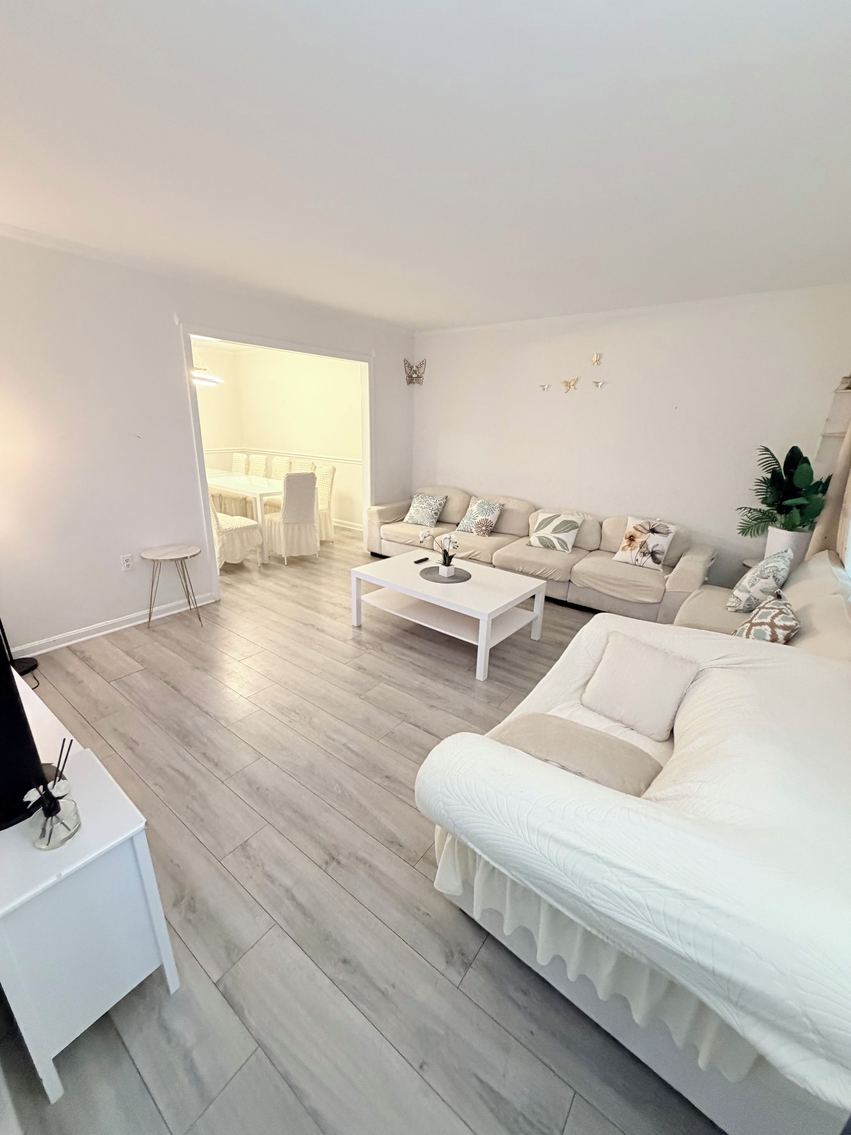Un salon moderne avec un ensemble de canapés blancs, une table basse et un coin repas visible à travers une porte ouverte.