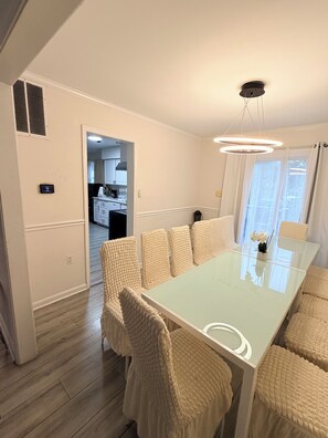 A modern dining area with a glass table and cushioned chairs, a ceiling light fixture, and a view into the kitchen.