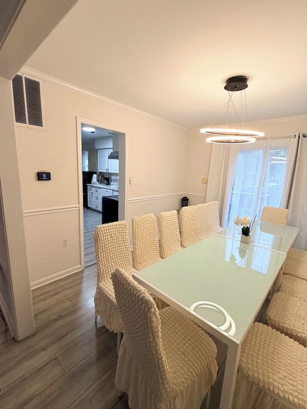 A modern dining area with a glass table and cushioned chairs, a ceiling light fixture, and a view into the kitchen. - Spacious 5-bedroom 11 beds house in awesome Silver Spring with AC (Silver Spring)