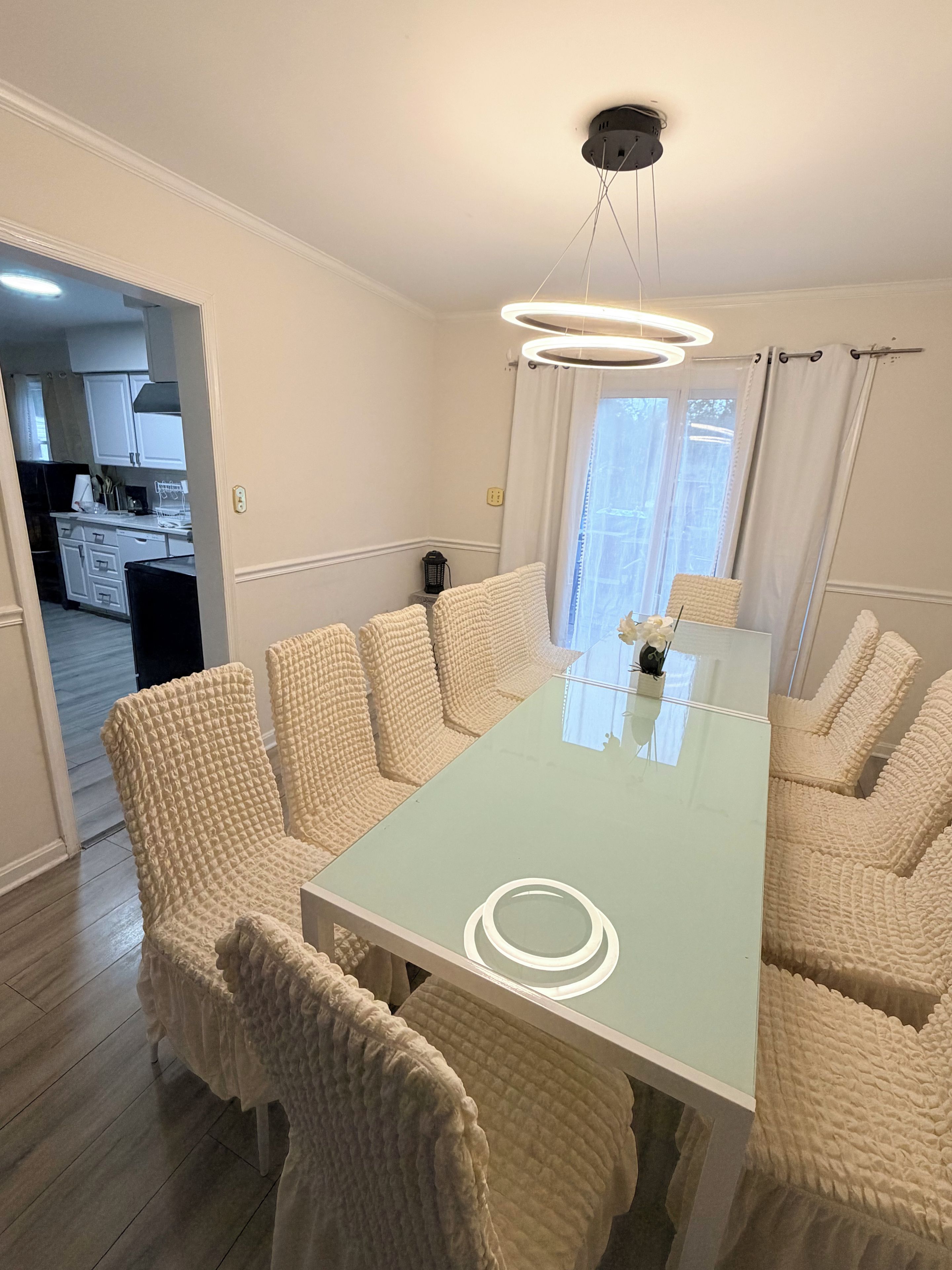A dining area with a glass-top table surrounded by six upholstered chairs, a ceiling light fixture, and a view of a kitchen through an open doorway.