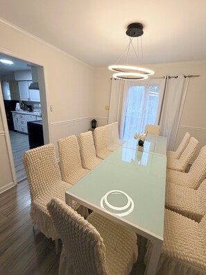 A dining area with a glass-top table surrounded by six upholstered chairs, a ceiling light fixture, and a view of a kitchen through an open doorway.