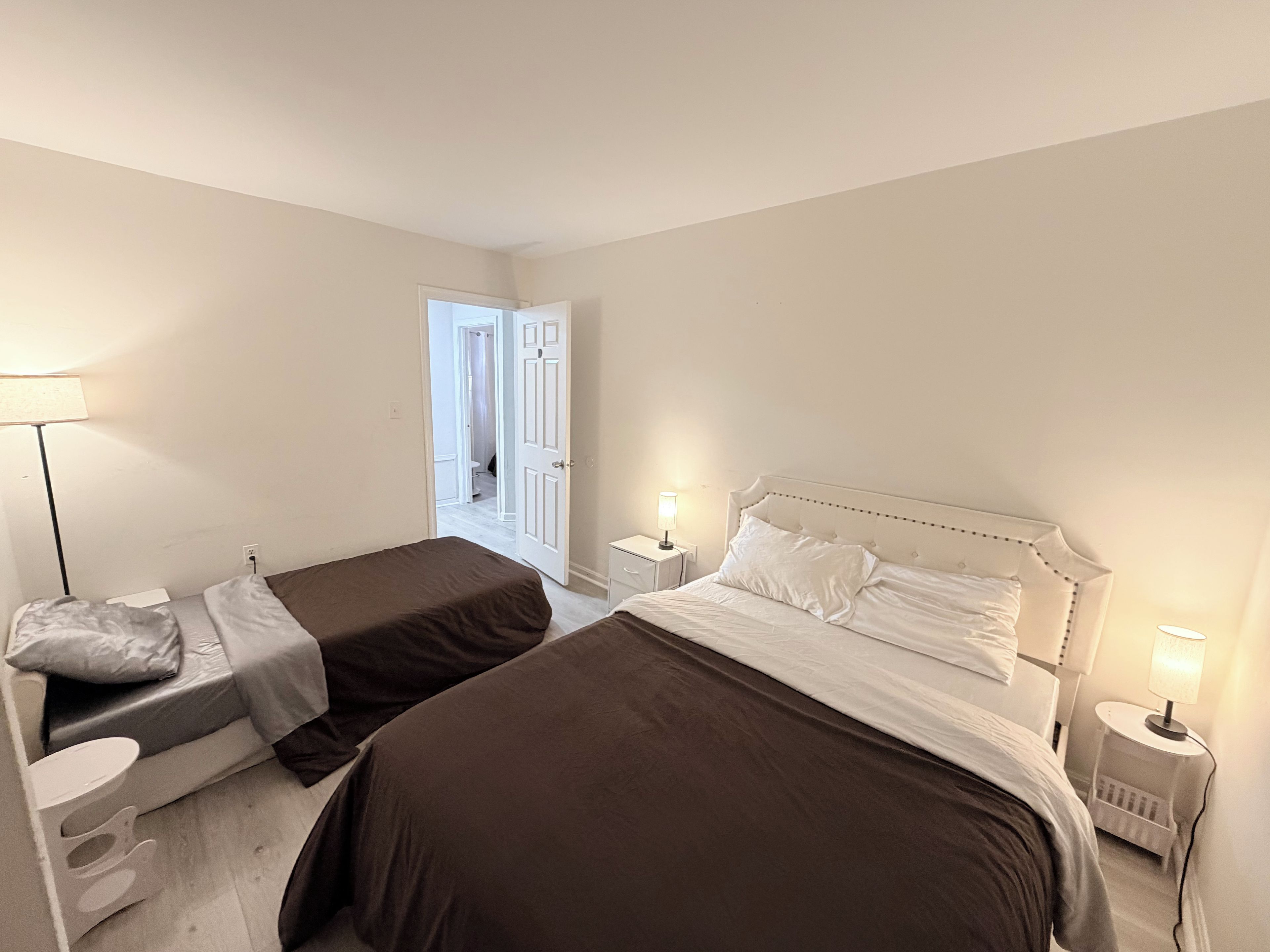 Un dormitorio con dos camas, una mesita de noche, una lámpara y una puerta que da a otra habitación.