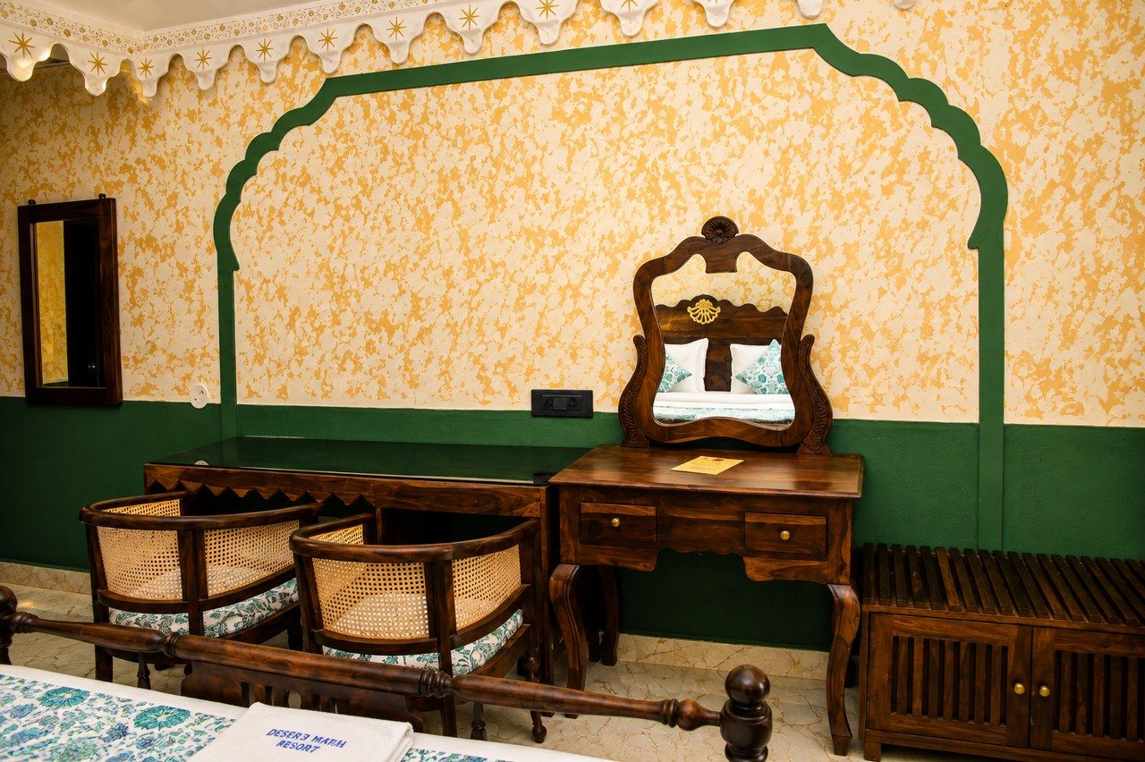 A room with a green and yellow patterned wall, a wooden desk with a mirror, wicker chairs, and a wooden bench.