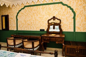 Une pièce avec un mur aux motifs verts et jaunes, un bureau en bois avec un miroir, des chaises en osier et un banc en bois.