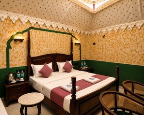 A four-poster bed with a wooden frame, a green and yellow patterned wallpaper, a white bedspread, and two pillows. There are two bedside tables with lamps, a round table, and two wicker chairs. - The Desert Mafia Resort & Camp (Jaisalmer)