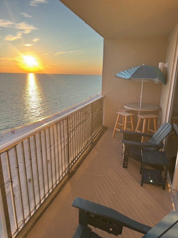A balcony with a view of the sunset over the sea, a table with chairs, and an umbrella. - Escape to Cloud 904 @ Tidewater Beach Resort on the water! (Panama City Beach)