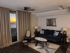 A living room with a dark blue sofa, two lamps, a starfish-patterned rug, a ceiling fan, and a view of the ocean through a sliding door. - Escape to Cloud 904 @ Tidewater Beach Resort on the water! (Panama City Beach)