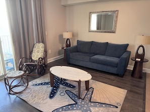 A living room with a blue sofa, a round coffee table, a patterned rug, a rocking chair, and a side table with a lamp. - Escape to Cloud 904 @ Tidewater Beach Resort on the water! (Panama City Beach)