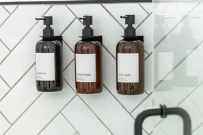Três frascos de produtos de higiene pessoal com bombas dosadoras, identificados como shampoo, condicionador e sabonete líquido, pendurados em uma parede revestida de azulejos.