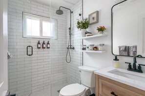 Ein modernes Badezimmer mit Dusche, Toilette und Waschbecken. Über der Dusche befindet sich ein Fenster, an der Wand ein Spiegel und auf Regalen verschiedene Pflegeprodukte.