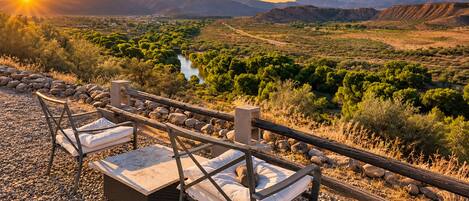 An outdoor seating area with two chairs and a table, overlooking a valley with a river and mountains in the distance.