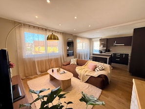 A modern living room with a sofa, coffee table, and a TV. There is a kitchen area with cabinets and a dining space. - Vrbo Property (Vesoul)
